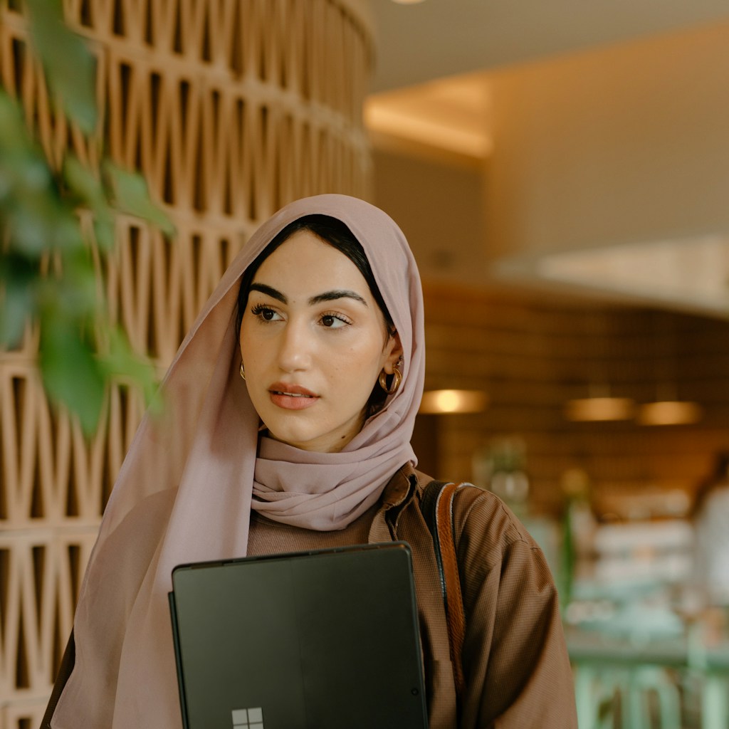 A Muslim professional woman in hijab, mid-day, discreetly feeling a prayer tap
